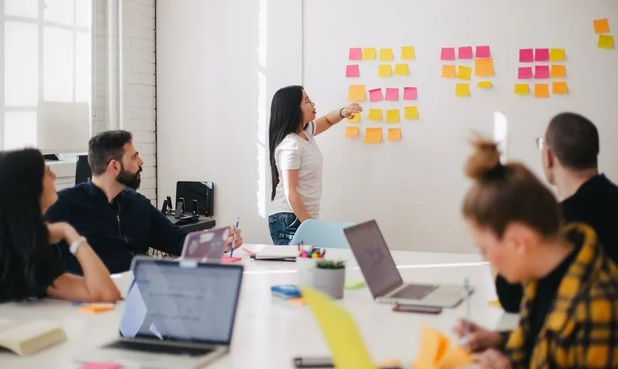 A woman presenting in a meeting. She is pointing towards a whiteboard covered in sticky notes